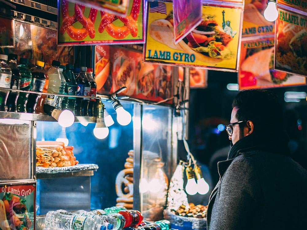 A person wearing glasses and a coat stands before a street food cart offering lamb gyros and pretzels during one of their day trips near Willamette Valley.