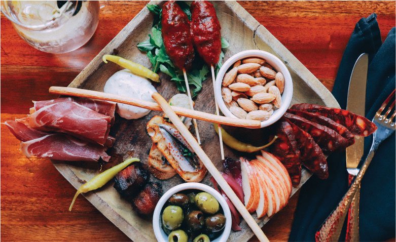 A wooden charcuterie board featuring skewers, cured meats, sliced apples, olives, almonds, and breadsticks, typical of restaurants in Yamhill County.