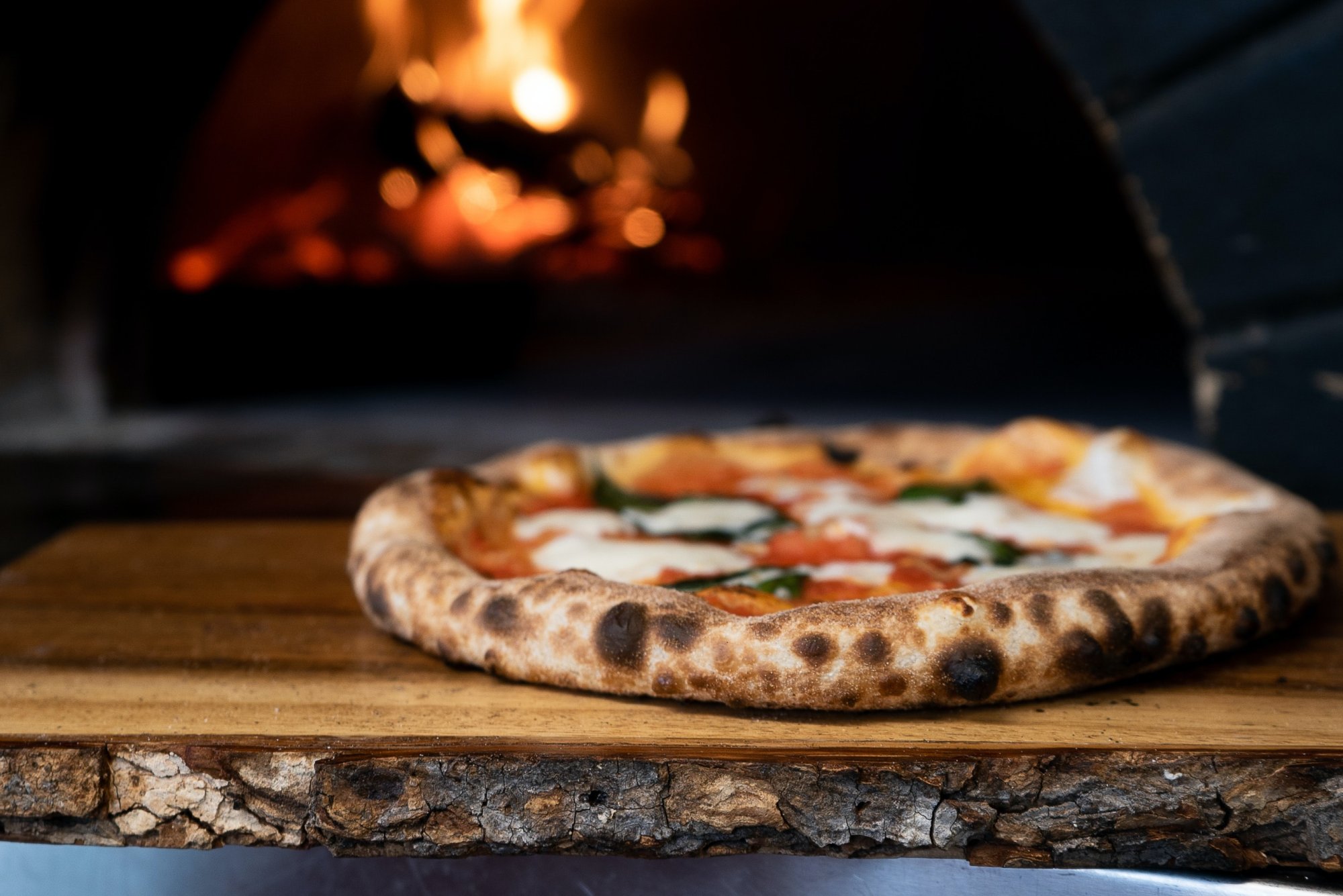 A wood-fired pizza with tomato sauce, mozzarella, and basil resting on a live-edge wooden board in front of a pizza oven at one of the restaurants in Yamhill County.