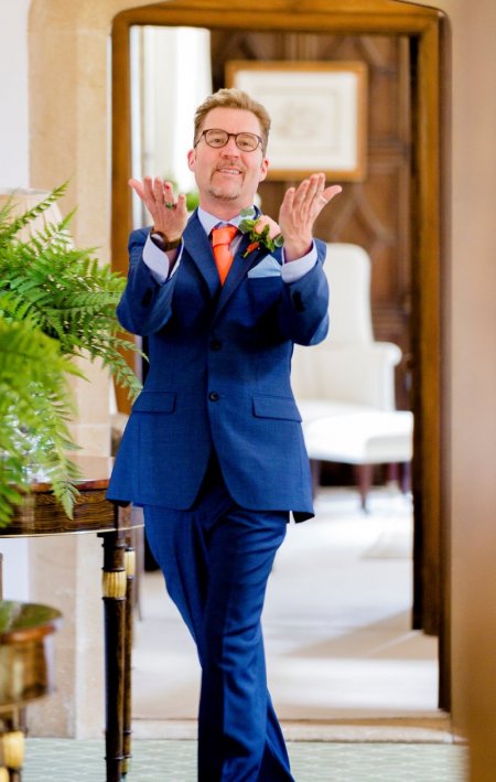A man in a blue suit with an orange tie smiles and gestures as he walks through a doorway at a bed and breakfast in Amity.