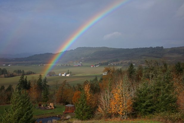 A vibrant rainbow arcs over a lush green valley and rolling hills during autumn.