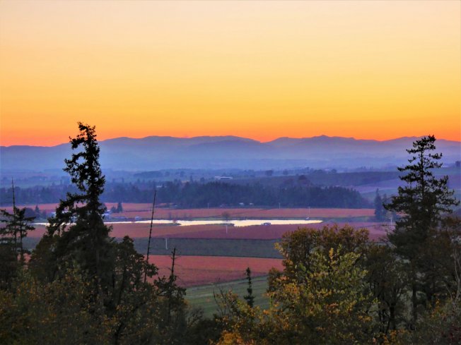 A sunset sky glows orange above silhouettes of distant mountains, a valley with a small lake, and foreground trees.